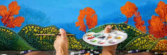 A person's hands are shown painting an autumn landscape featuring vibrant orange trees and rolling hills with yellow flowers on a canvas.