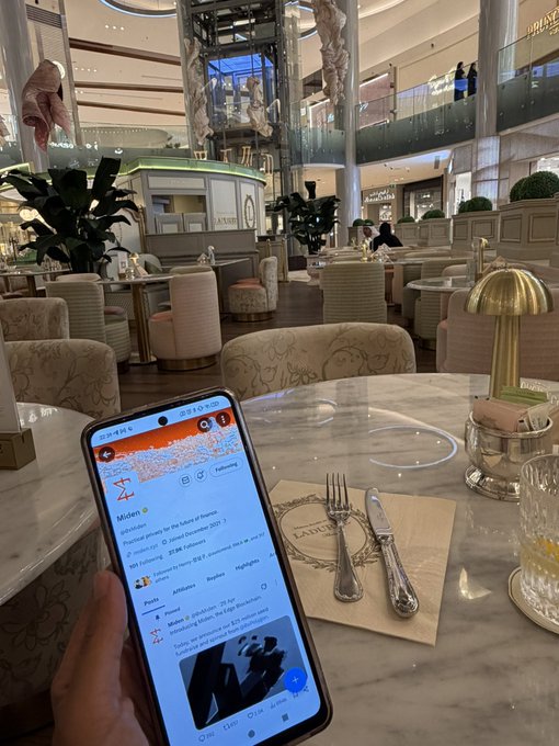 A first-person view of a hand holding a smartphone displaying a social media app in an elegant, upscale shopping mall or hotel lobby with plush chairs, dining tables, and a glass elevator.