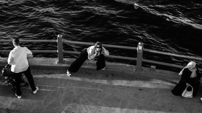 A black and white photograph taken from above shows people on a waterfront promenade, including one person leaning on the railing taking a photo.