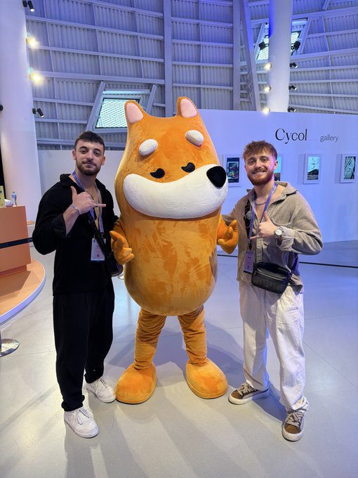 Two attendees pose with a large Shiba Inu mascot while flashing hand signs at what appears to be the "Cycol gallery" exhibit, likely sharing a cheerful "GM" (good morning) greeting to the crypto/Web3 community.