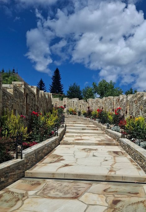 A scenic outdoor stone staircase flanked by stone walls and colorful flower planters leads upward under a bright blue sky with scattered white clouds.