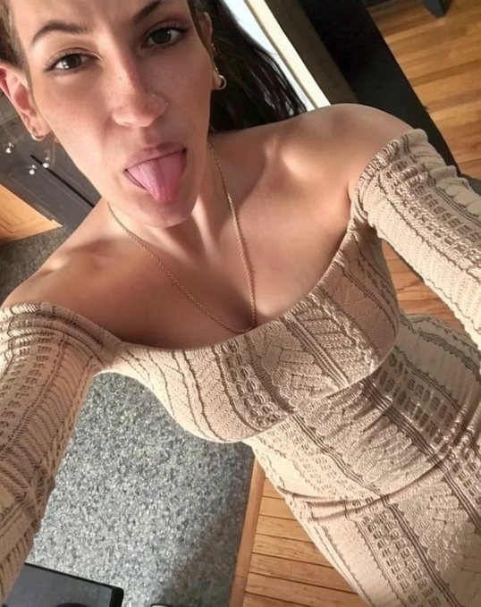 A selfie of a woman wearing a beige off-the-shoulder ribbed dress and a necklace, sticking her tongue out at the camera.
