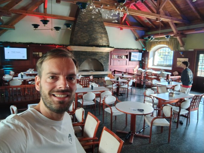 A smiling man takes a selfie inside a spacious, empty restaurant featuring a vaulted wooden ceiling, a central stone fireplace, and numerous set tables.
