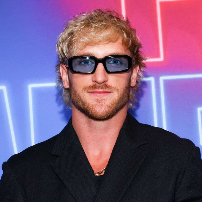 A man with curly blond hair and a beard wearing dark rectangular sunglasses and a black blazer stands in front of a neon-lit backdrop with blue and pink lighting. The image relates to the accompanying post about YouTuber Logan Paul's NFT investment, which he purchased for $635,000 in 2021 but has since plummeted in value to just $96.