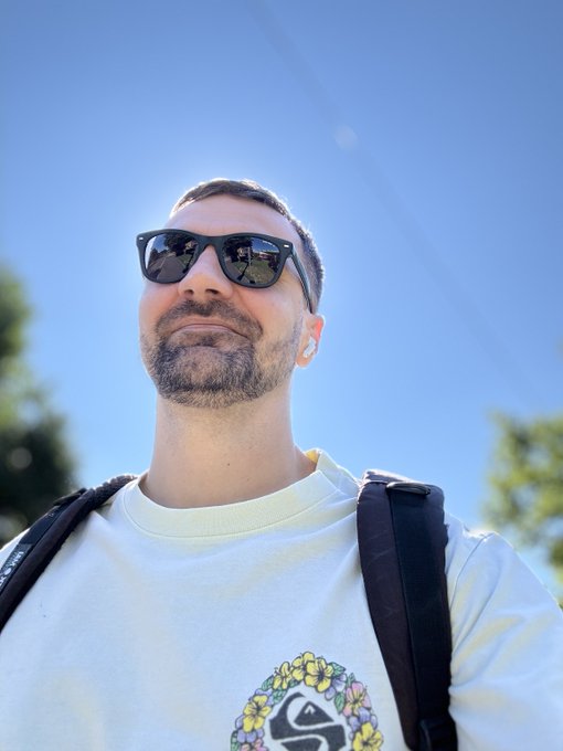 A man wearing sunglasses and earbuds poses for a low-angle selfie against a clear blue sky, wearing a light-colored t-shirt with a graphic design and a backpack.