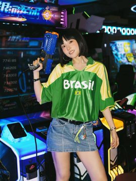 A young woman wearing a green and yellow Brazil soccer jersey and denim shorts, holding a blue toy gun in what appears to be an arcade setting.