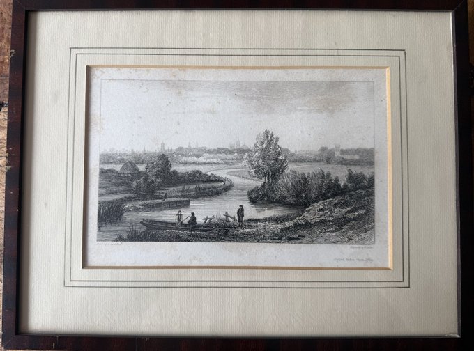 Framed antique engraving/etching depicting a pastoral riverside landscape with small figures by the water, trees, and a distant town, presented in a dark wooden frame with cream matting.