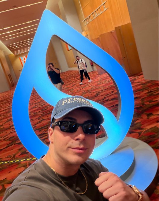 A man wearing a denim 'present' cap and sunglasses takes a selfie in front of a large blue flame or water drop-shaped logo installation at what appears to be a conference venue, with other people visible in the background.