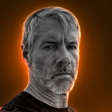 A black-and-white portrait of a gray-haired, bearded man (appearing to be Michael Saylor) is set against a glowing orange backdrop, visually evoking the heat of scrutiny around his Bitcoin strategy. The image reinforces the post's theme of Saylor's Strategy company facing trouble as its mNAV metric reveals the market undervaluing its Bitcoin holdings.