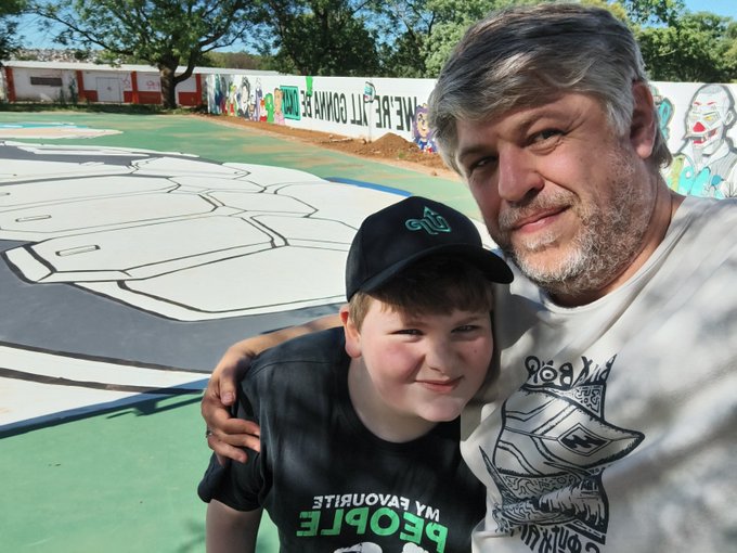 A man and a young boy pose for a selfie in front of a colorful outdoor basketball court mural, with the boy wearing a black cap and a "MY FAVOURITE PEOPLE" t-shirt. The caption "GM to everyone who celebrated Taco Tezo Tuesday" appears unrelated to the image content.