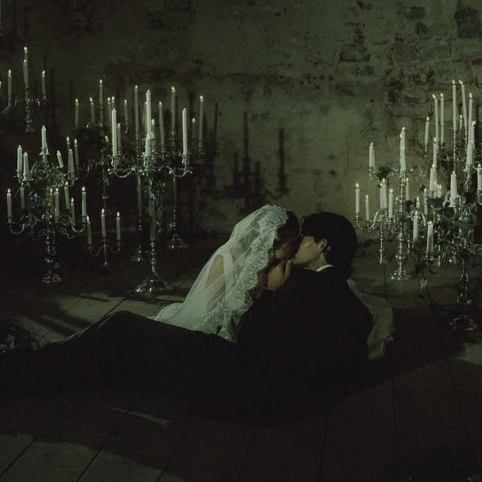 A bride and groom share a kiss in a dimly lit, atmospheric room surrounded by tall candelabras casting dramatic shadows.