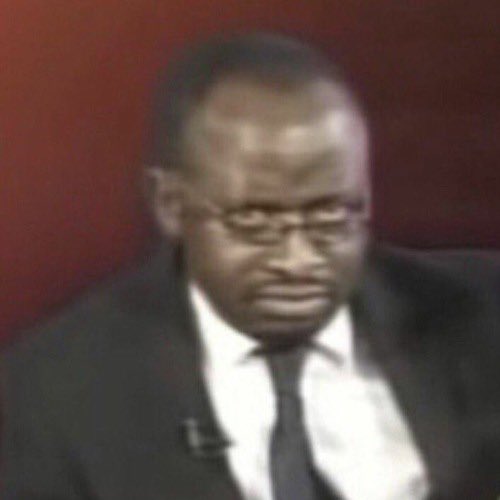 A low-quality, blurry image shows a man wearing glasses, a dark suit, white shirt, and tie against a dark red background.