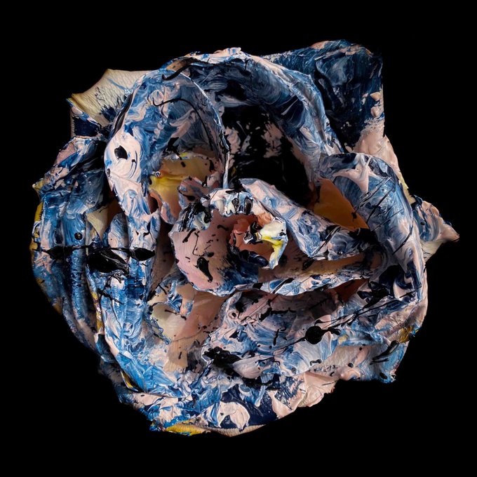A textured, sculptural object resembling a rose, heavily painted with thick strokes of blue, white, pink, and yellow against a solid black backdrop.