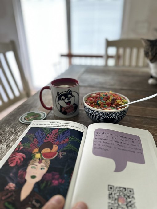 A cozy breakfast scene featuring a colorful bowl of Froot Loops and a Husky-themed mug sit beside an open book showcasing vibrant Frida Kahlo artwork, with a curious cat peeking in from the right. The page displays the inspirational quote "How do you live when you are told you are dying? It is important to adjust your lens to move forward. Mental discipline is a choice in overcoming any challenge," paired with a QR code for accessing the full "Fire within" reading.