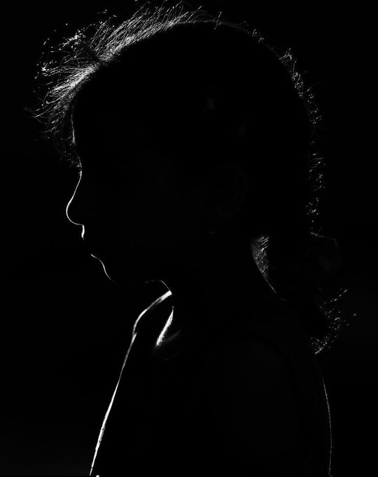 A high-contrast black and white profile silhouette of a person with long hair, illuminated by a thin sliver of light against a dark background.