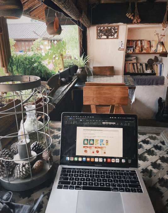 A MacBook displaying a webpage sits on a woven mat in a rustic, plant-filled open-air workspace, surrounded by wooden furniture, a bookshelf, and hanging decorations, capturing the essence of remote work from a scenic travel destination.