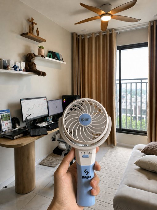 A hand holding a blue and white Bitget-branded portable fan in the foreground, with a home office setup featuring dual monitors, a ceiling fan, curtains, and a balcony view in the background.