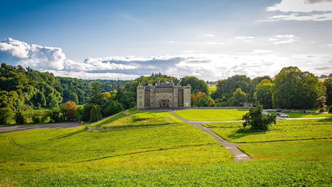 A picturesque landscape showing a stone castle or manor house surrounded by lush green trees and expansive grassy fields under a bright, partly cloudy sky.