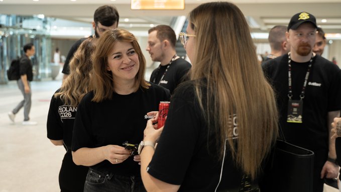 Two women chat at the Solana Summit Serbia, one wearing a black t-shirt with "SOLAN 2026" visible on the side while holding a phone, as another woman in sunglasses and a black "SOLANA" t-shirt listens, with event staff in lanyards standing nearby in the busy conference venue.