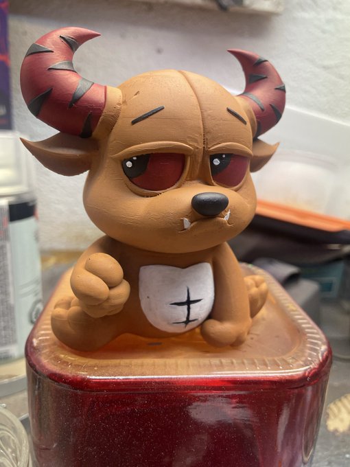 A hand-painted, custom-crafted vinyl figure of a baby bullbear sits atop a red ceramic base, featuring distinctive red horns, droopy red eyes, a small fanged mouth, and a white belly patch with a black stitched X design. The post playfully announces the figure as a "baby bullbear brought to life," perfectly matching the small, chibi-style creature shown in mid-creation.