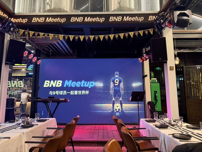A view of the BNB Meetup event stage featuring a large screen displaying a football player wearing jersey number 9 with the text "BNB Meetup" and "与9号球员一起看世界杯" (Watch the World Cup with Player #9), set up with dining tables in the foreground for guests to enjoy food and drinks while watching the sports broadcast.