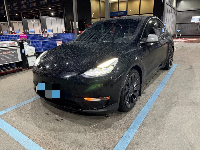 A black Tesla Model Y, still covered in water droplets, is parked at a charging station with its license plate blurred and blue parking lines visible on the ground. The owner's comment about being "still dissatisfied" with the car suggests frustration or unmet expectations despite its sleek appearance.