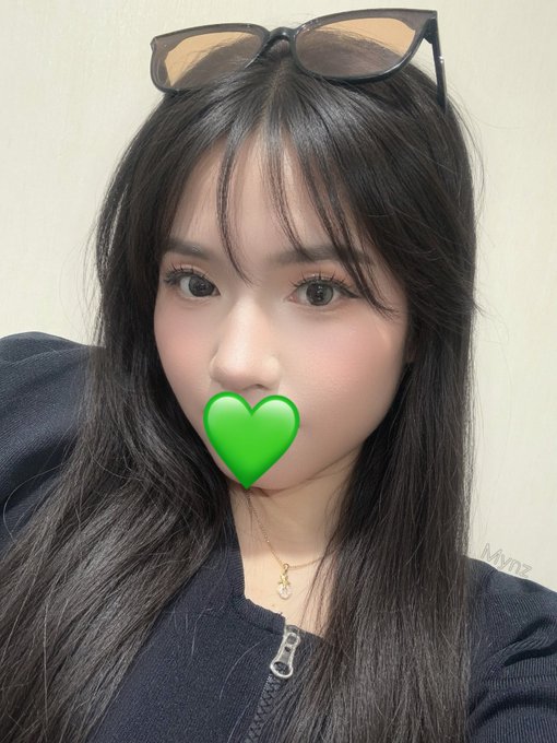 Close-up selfie of a young woman with long dark hair, sunglasses on her head, wearing a black zip-up jacket and a gold necklace, with a green heart emoji covering her mouth.