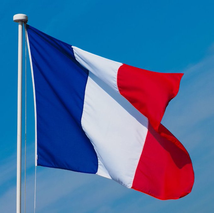 A French flag with blue, white, and red stripes waves against a clear blue sky on a flagpole.