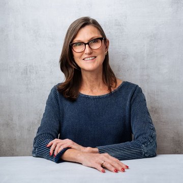 A woman with shoulder-length brown hair and glasses, wearing a navy blue textured sweater, smiles warmly with her arms resting on a table. She appears to be Cathie Wood, the ARK Invest CEO, pictured in a professional headshot that accompanies the article about her bullish outlook on Bitcoin's breakout relative to gold.