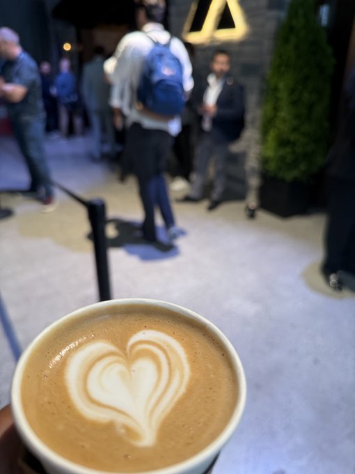 A first-person view of a latte with heart-shaped latte art, held up against the blurred background of an outdoor gathering area where people are standing around.