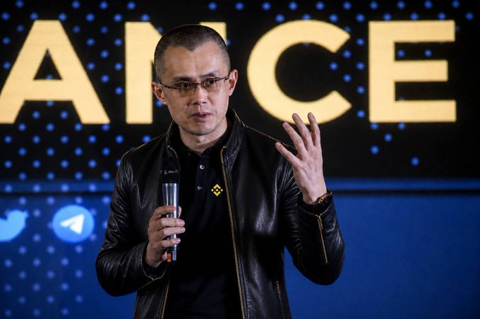 A man with short hair and glasses, wearing a black leather jacket featuring a Binance logo, speaks into a microphone at a Binance event with the partial word "ANCE" visible on a large screen behind him. The image ties to the post's playful reference to CZ's "world-changing JACKET," highlighting his signature style and the crypto exchange's branding prominently displayed on his attire.