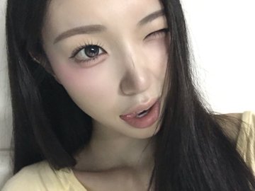 A close-up selfie of a young woman with long dark hair, pale skin, and a subtle winged eyeliner look, wearing a yellow top.
