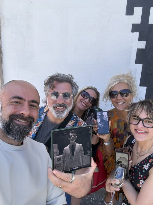 The image features a group of five people posing together for a photo, with one person holding up an album or CD cover featuring a picture of a man wearing sunglasses. The group appears to be enjoying their time together as they share this moment.