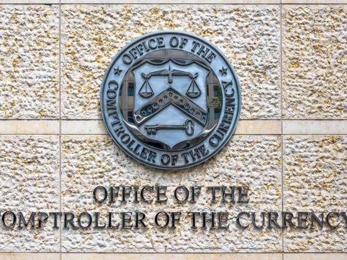 A stone building facade displays a metal seal and engraved text reading "OFFICE OF THE COMPTROLLER OF THE CURRENCY," featuring a shield emblem with scales of justice and a key.
