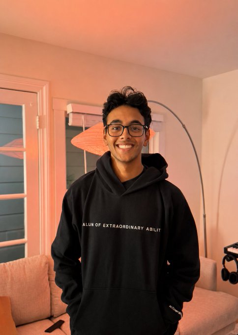 A smiling young man wearing glasses and a black hoodie featuring the text "ALIN OF EXTRAORDINARY ABILIT" poses in a warmly lit room with a modern floor lamp and sofa. The image appears to accompany the post's casual announcement about GPT usage limits, fitting its laid-back, conversational tone.