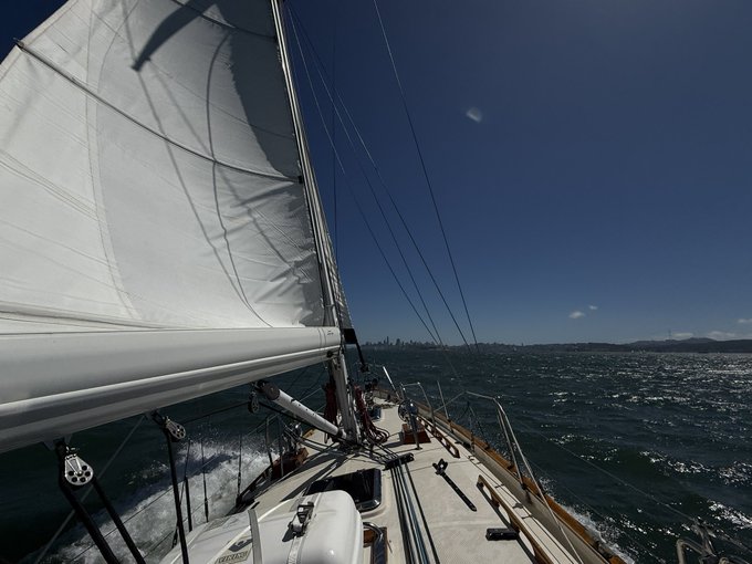 A first-person view from the helm of a sailing yacht shows a white sail billowing overhead and the bow cutting through choppy blue waters, with a distant city skyline (likely San Francisco) visible on the horizon under a clear sky. The image perfectly complements the "Back at the (literal) helm" caption by placing the viewer directly in the captain's perspective at the ship's wheel, navigating toward the iconic skyline.