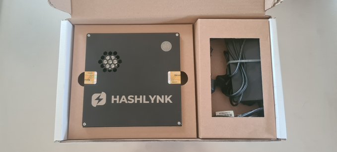 An open cardboard box containing a black "HASHLYNK" branded hardware device (likely a crypto miner) alongside its power supply and cables.