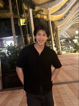 A young man wearing a black short-sleeved button-down shirt poses for a photo at night in front of a brightly lit building with palm trees.