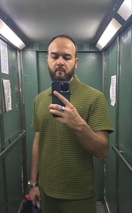 A man with a beard and eyes closed taking a mirror selfie in a narrow space with green metal walls, wearing a textured olive-green knit polo shirt and matching pants, holding a dark blue smartphone and a rolling suitcase.