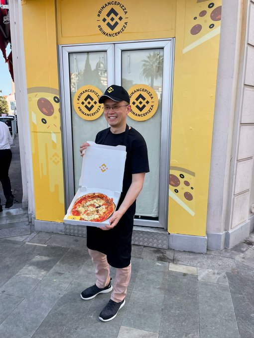 A smiling man wearing a black Binance Pizza cap and t-shirt standing in front of a yellow Binance Pizza storefront, holding an open pizza box with a baked pizza inside.