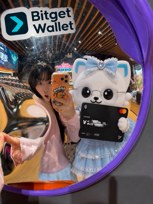 The image features a woman taking a selfie with a cute white stuffed animal, possibly a teddy bear or a cat-like creature, while holding a credit card. The scene takes place inside a store, as indicated by the presence of a mirror in the background. The woman is standing next to the mirror and appears to be capturing this moment on her phone. The image also includes text that reads "Bitget Wallet CN" and "@bitget wallet CN", which might be related to the credit card used for payment or the store's name, respectively.