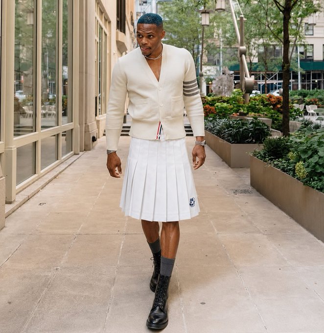 A man with blue hair walks down a city sidewalk wearing a cream cardigan, a white pleated tennis skirt, gray socks, and black combat boots.