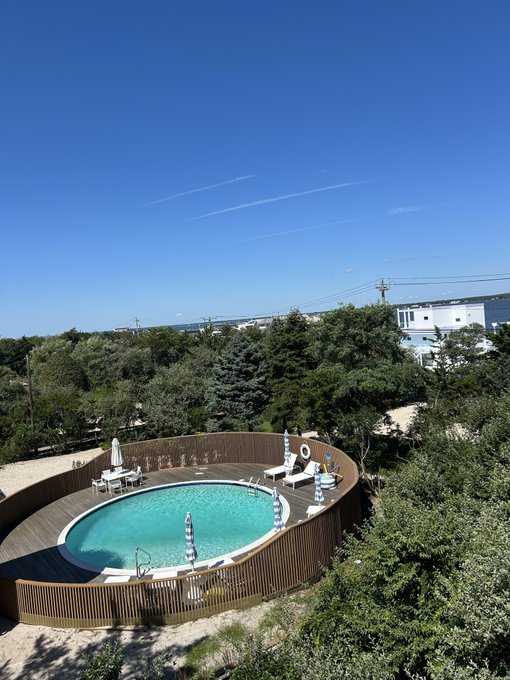 A round above-ground swimming pool with a wooden deck and striped umbrellas sits nestled among trees on a sunny day, with a clear blue sky overhead. The "gm mfers" (good morning) caption reflects the bright, cheerful morning vibe of this serene backyard scene.