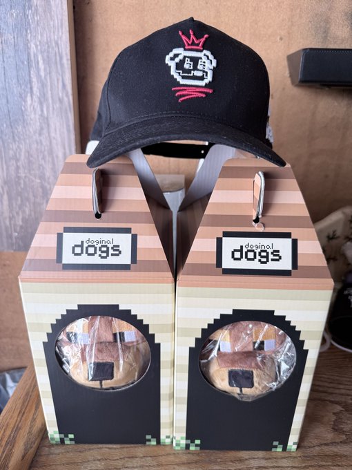 A black baseball cap with pixel-art 'LB' logo and pink crown embroidery sits on top of two 'dosinol dogs' boxes with pixel-art designs and windows showing bread contents.