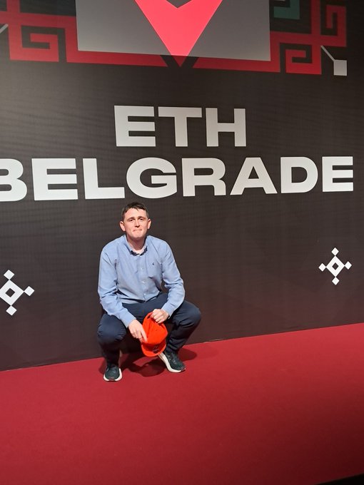 A man wearing a blue shirt crouches on a red carpet while holding an orange cap, posing in front of an 'ETH BELGRADE' sign.