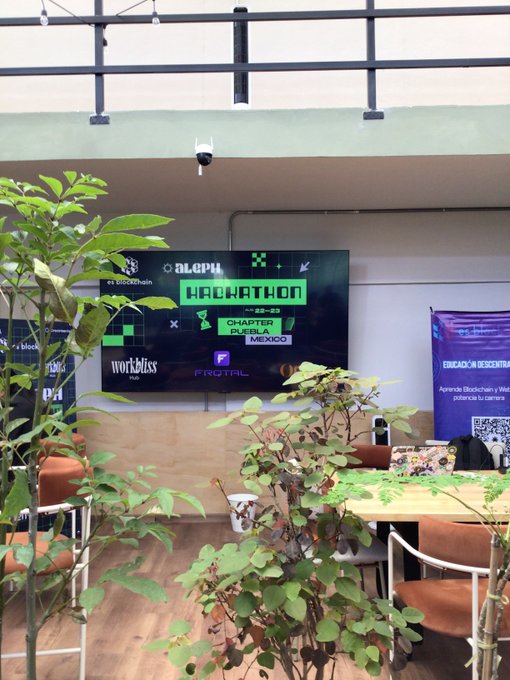 An indoor photo of a space decorated for an Aleph Hackathon on Chapter Puebla Mexico, featuring banners, tables, chairs, and potted plants.