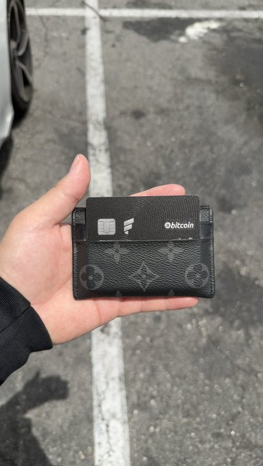 The image features a person holding up a black wallet with a Bitcoin logo, which has been placed inside of an ATM machine. The wallet appears to be made from leather or a similar material, and it contains a credit card. The text "Best credit card out there matches my card holder @fold_app #bitcoin" is present in the image, indicating that this wallet is specifically designed for Bitcoin users.