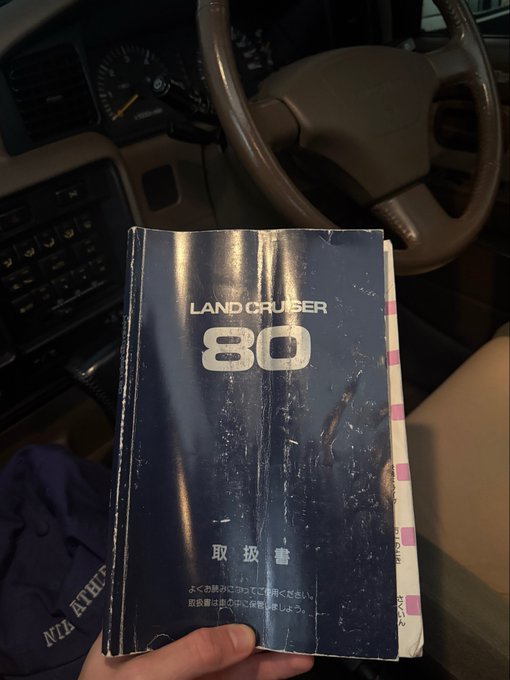 A hand holds a well-worn Japanese owner's manual for a "LAND CRUISER 80" inside the cabin of the vehicle, with the steering wheel and dashboard visible in the background, fitting the post's reference to a "land cruiser delivered."