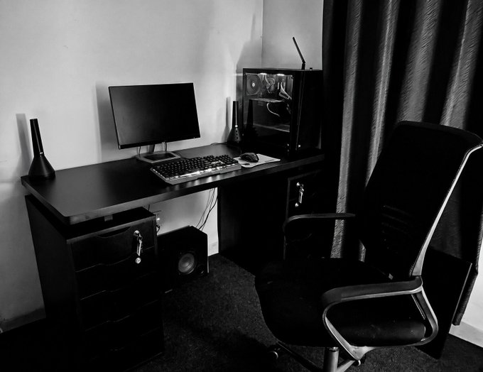 A black and white photo of a minimalist home office setup featuring a black desk, monitor, keyboard, PC tower, speaker, and an office chair.