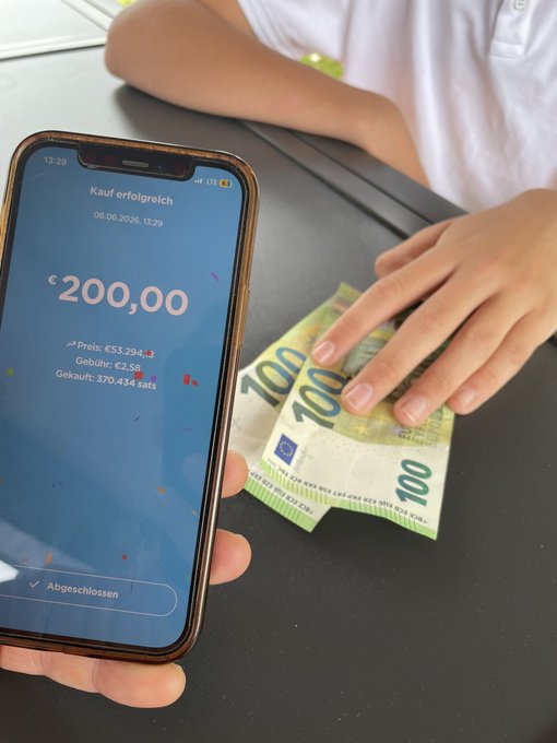In this image, a person's hand is holding a black iPhone displaying an app where they are sending money over Bitcoin or cryptocurrency. The person sitting at a table also has a stack of Euro banknotes nearby. There is no visible text present, but from these elements, it seems like the person might be transferring funds to someone using Bitcoin or another cryptocurrency instead of traditional payment methods.